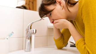 Junge Frau im gelben Pullover stützt sich auf Waschbecken ab und hält sich vor Übelkeit die Hand vor den Mund