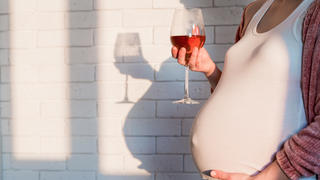 Schwangere Frau hält ein Glas Rotwein in der einen Hand und hält mit der anderen Hand ihren Babybauch fest