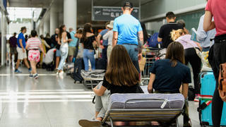 29.07.2018, Bayern, München: Fluggäste sitzen auf einem Klappbett in einer langen Schlange an einem Lufthansa-Schalter im Terminal 2 am Flughafen in München. Nach einem Polizeieinsatz am Vortag mit über 200 ausgefallenen Flügen sind die Konsequenzen am Terminal 2 am Flughafen München noch immer spürbar. (zu dpa "Weiter Ausfälle und Verspätungen am Flughafen München" vom 29.07.2018) Foto: Matthias Balk/dpa +++ dpa-Bildfunk +++