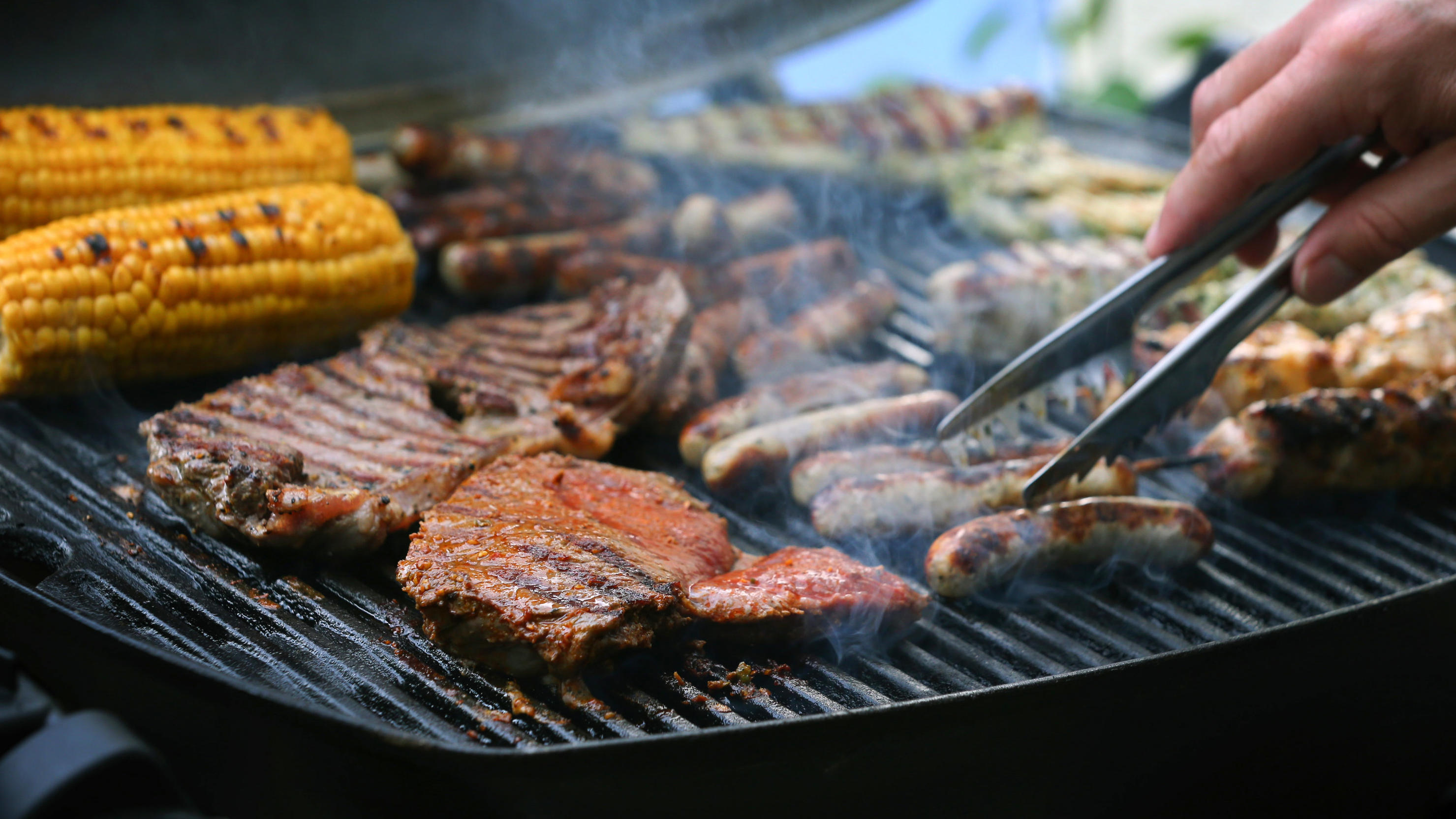 Maiskolben, Fleischspieße, Bratwürste und Steaks liegen auf dem Rost eines Gasgrills. 
