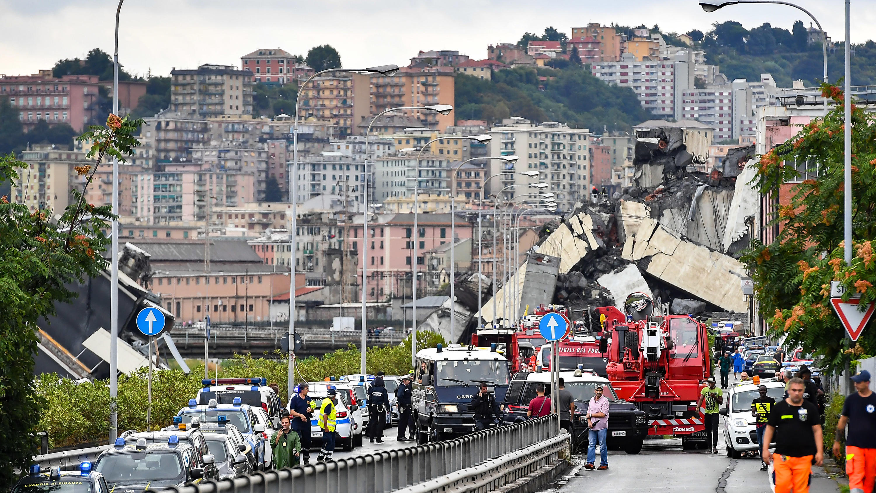 14.08.2018, Italien, Genua: Feuerwehrwagen und Polizeiautos sind vor der teilweise eingestürzten Autobahnbrücke Ponte Morandi zu sehen. Beim Einsturz der Brücke sind mindestens elf Menschen ums Leben gekommen. Das berichtete die Nachrichtenagentur An
