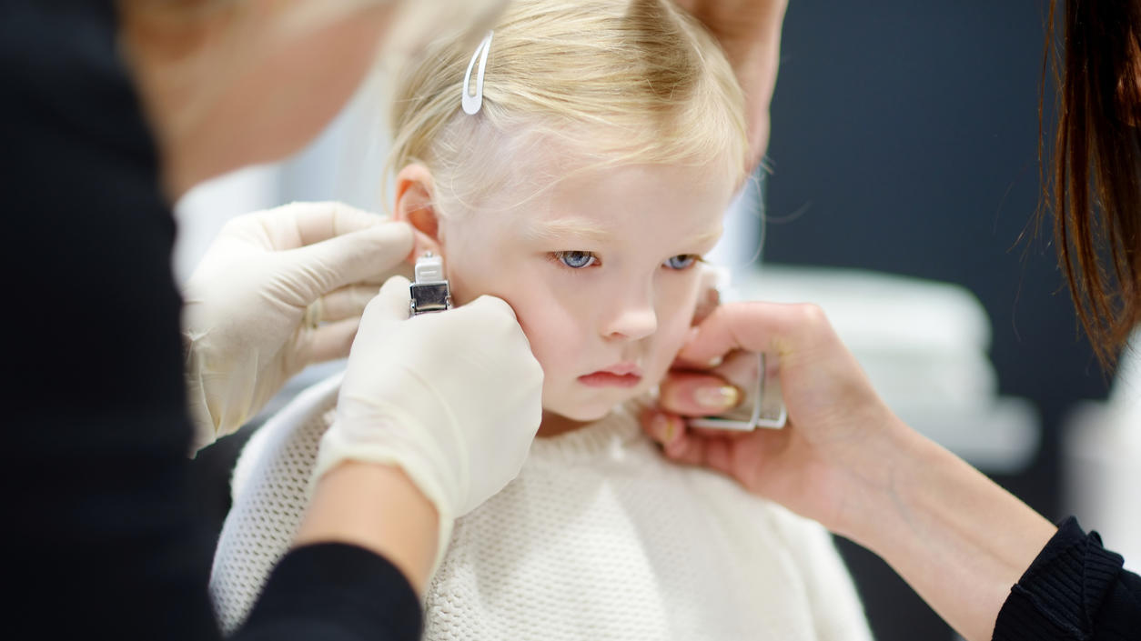 Adorable little girl having ear piercing process with special equipment in beauty center by medical worker
