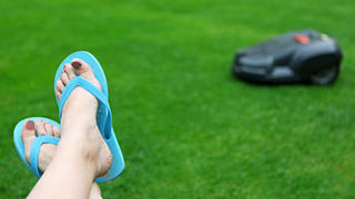 This is an image of an automatic lawn mower mowing the grass while a person is resting. Women's feet with flip flops are seen in the foreground, while the robot mower is in the background against green grass background.