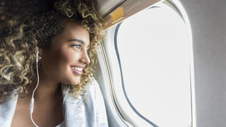 Confident young mixed race woman smiles while looking through window on aircraft. She is wearing earbuds.