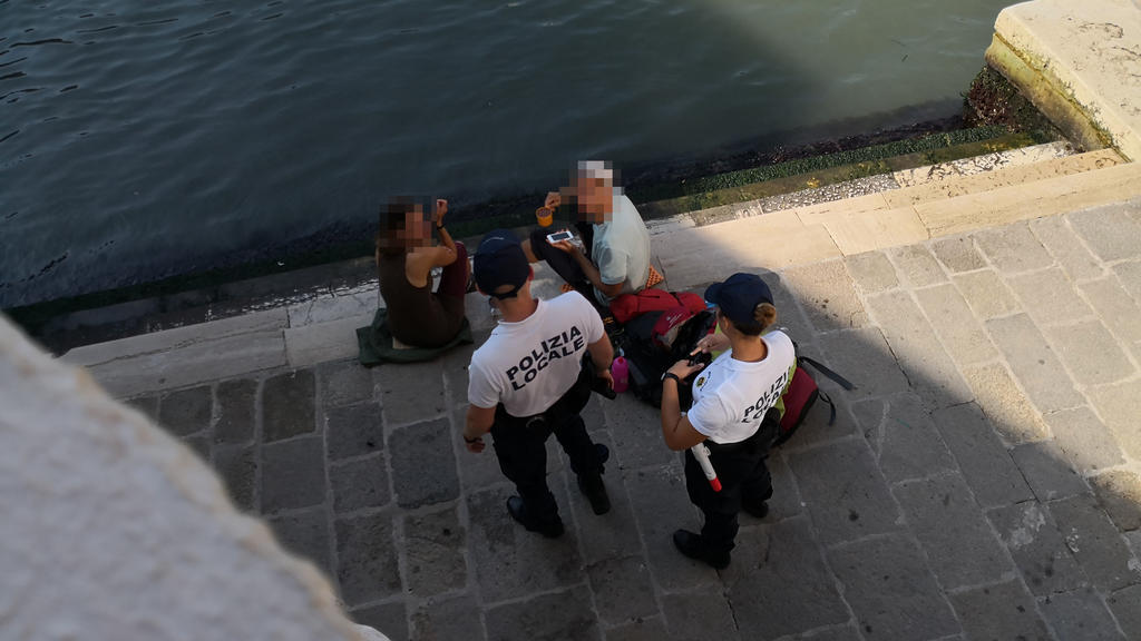HANDOUT - 19.07.2019, Italien, Venedig: Zwei deutsche Touristen werden von zwei Polizisten an der Treppe der Rialto-Brücke angesprochen, da sie sich mit einem kleinen Kaffeekocher Kaffee auf dem Gehweg zubereitet haben. (zu dpa «Deutsche Touristen ko