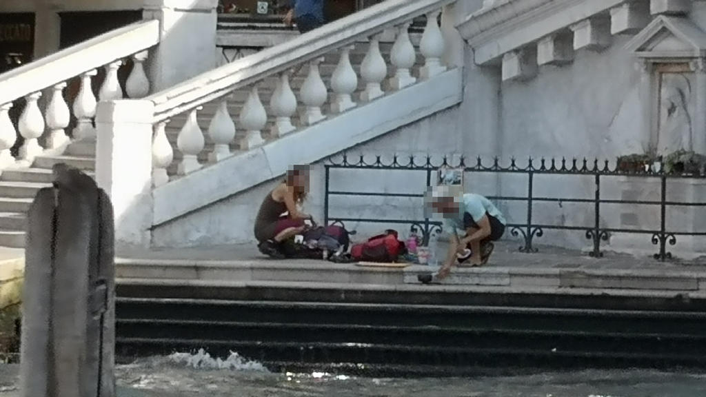 HANDOUT - 19.07.2019, Italien, Venedig: Zwei deutsche Touristen sitzen mit ihrem Gepäck an der Treppe der Rialto-Brücke und kochen sich mit einem kleinen Kaffeekocher einen Kaffee auf dem Gehweg. (zu dpa "Deutsche Touristen kochen Kaffee in Venedig -