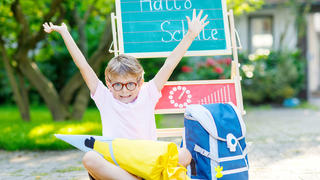 Happy little kid boy with glasses sitting by desk and backpack or satchel. Schoolkid with traditional German school bag called Schultuete on his first day to school. Hello school in German language
