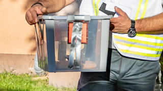 30.08.2019, Nordrhein-Westfalen, Herne: Reptilienexperte Roland Byner trägt die entwischte Monokelkobra in einem Behälter aus einem Hauseingang. Am 25. August war im Treppenhaus eines Mehrfamilienhauses eine hochgiftige Monokelkobra mit einer geschätzten Länge von mindestens 1,40 Metern gesichtet worden. Mehrere Mehrfamilienhäuser wurden evakuiert. Das Tier wurde jetzt gefangen. Foto: Marcel Kusch/dpa +++ dpa-Bildfunk +++