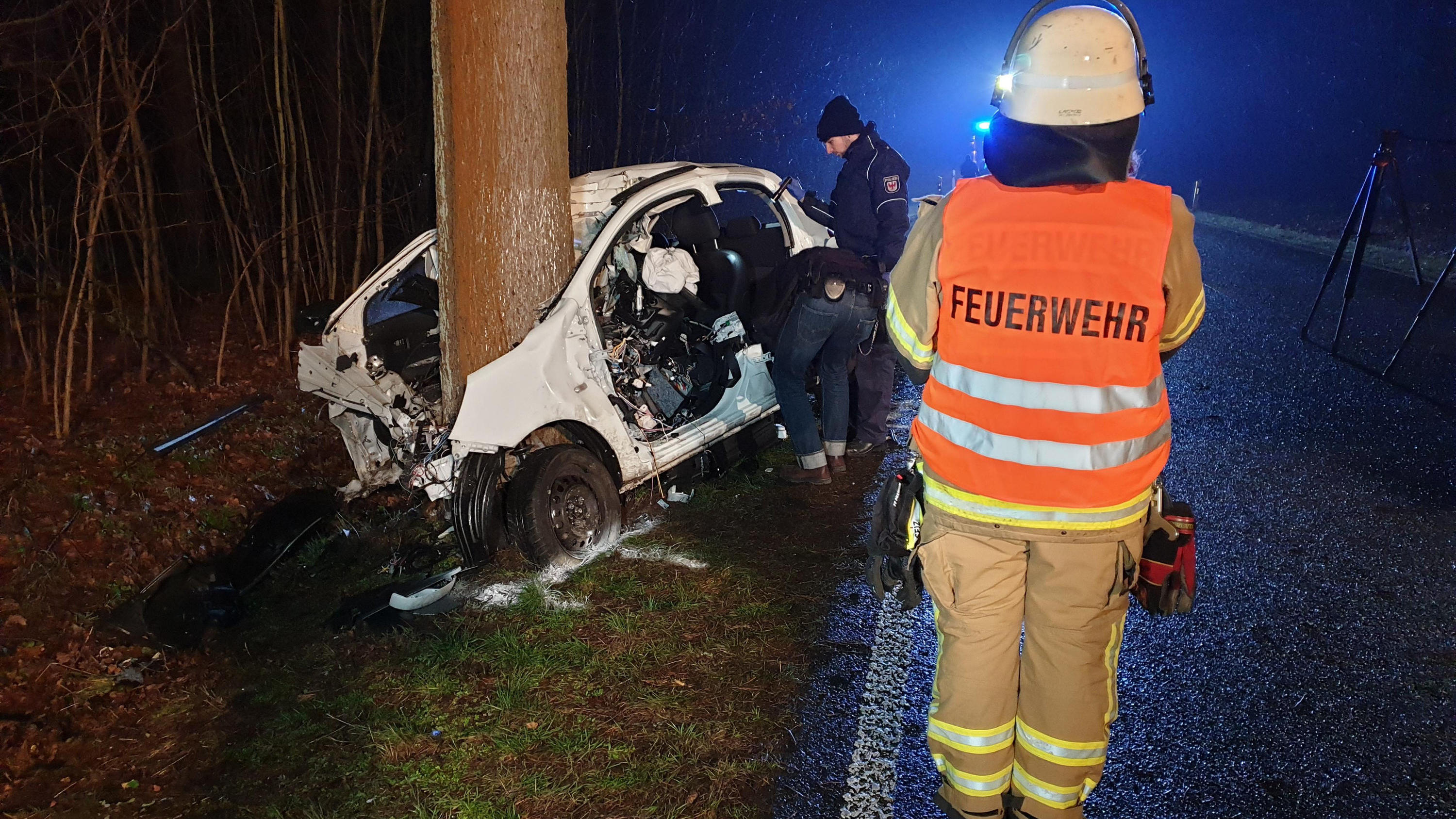 Unfall in Brandenburg: Zwei Polizisten und ein Feuerwehrmann stehen neben dem stark beschädigten Auto.