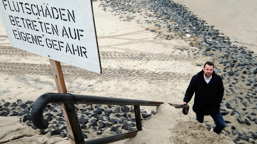 13.02.2020, Niedersachsen, Wangerooge: Ein Schild mit der Aufschrift "Flutschäden, Betreten auf eigene Gefahr" ist am Strand zu sehen. Bürgermeister Marcel Fangohr (parteilos) steht im Hintergrund an der Abbruchkante des Strandes. Die Sturmflut der l