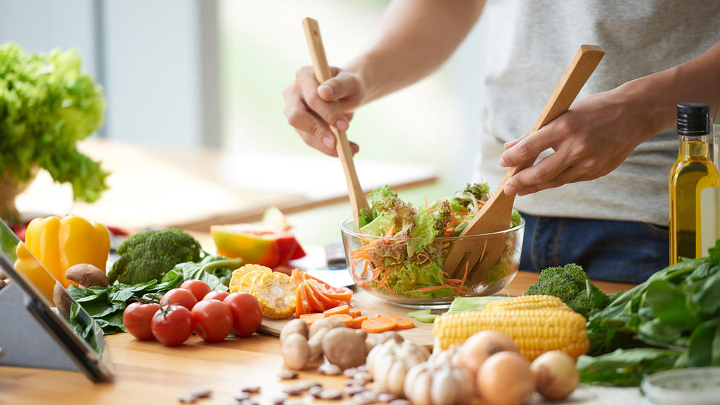 Ein Mann mixt in einer Salatschüssel seinen Salat, um ihn herum liegen verschiedene Gemüsesorten.