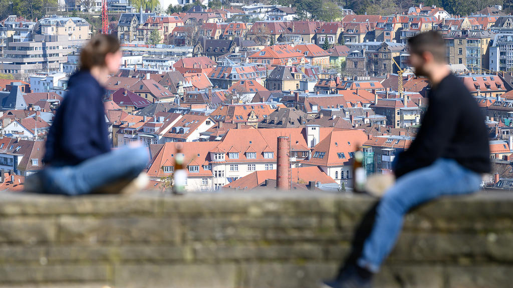05.04.2020, Baden-Württemberg, Stuttgart: Eine junge Frau und ein junger Mann sitzen im Abstand von etwa eineinhalb Metern voneinander entfernt auf einer Mauer auf der Karlshöhe. Im Hintergrund sind die Dächer der Innenstadt zu sehen. Derzeit herrsch