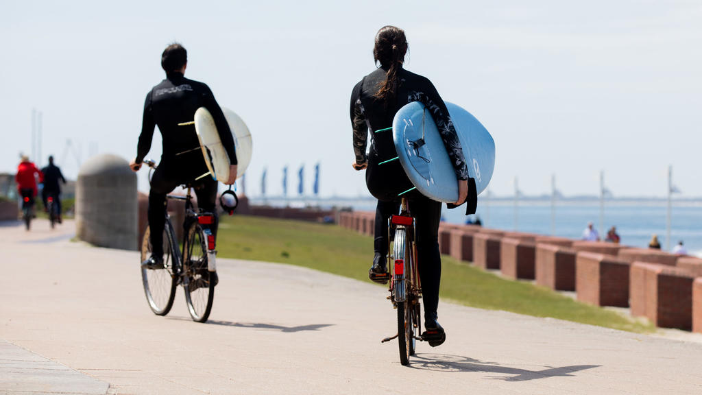 26.05.2020, Niedersachsen, Norderney: Zwei Surfer auf Fahrrädern fahren mit ihren Surfbrettern auf der Promenade am Meer entlang. Foto: Rolf Vennenbernd/dpa +++ dpa-Bildfunk +++