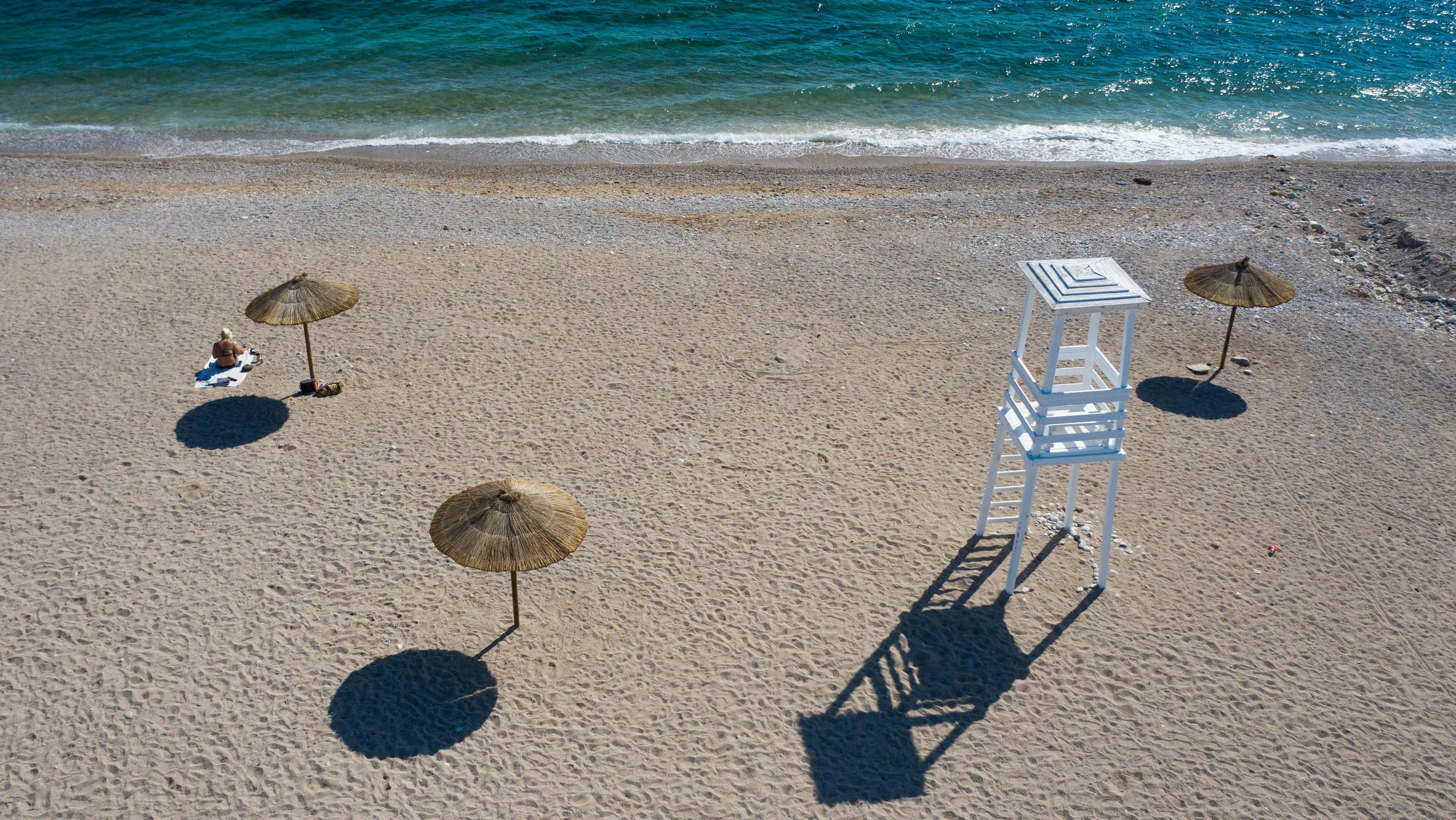 30.05.2020, Griechenland, Athen: Eine Frau sitzt an einem Strand mit Blauer Flagge südlich von Athen neben einem Sonnenschirm in der Sonne. Athen hat angekündigt, den Tourismus aus dem Ausland neu starten zu wollen. Heute soll der griechische Pandemi