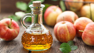 Eine durchsichtige Flasche mit Apfelessig steht vor losen Äpfeln auf einem Holztisch