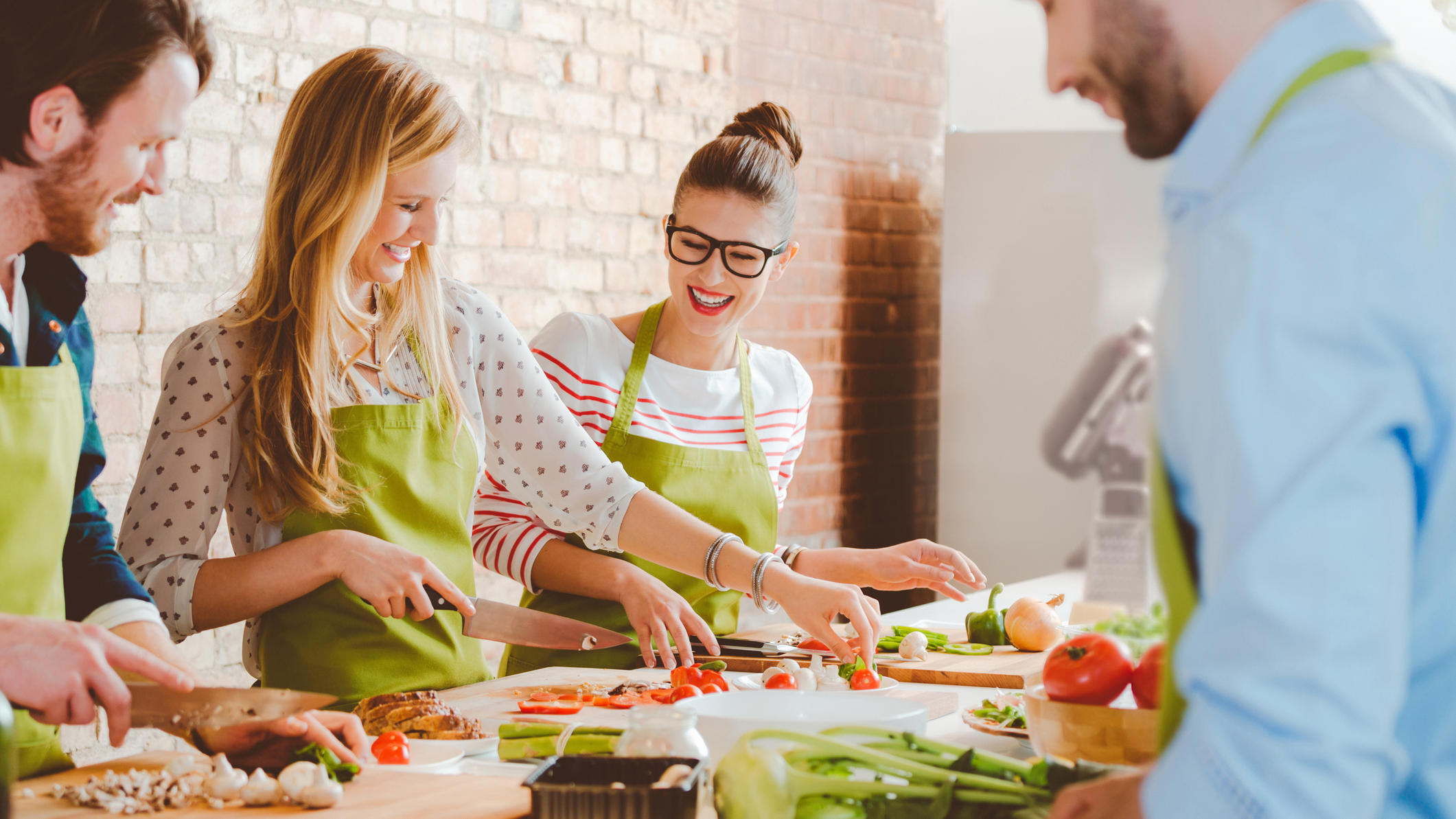 Group of people wearing aprons taking part in cooking class, preparing food, slicing vegetables, talking, laughing.