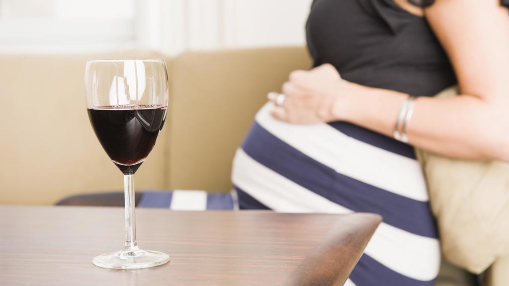 Ein Glas Rotwein auf einem Tisch, schwangere Frau im Hintergrund.
