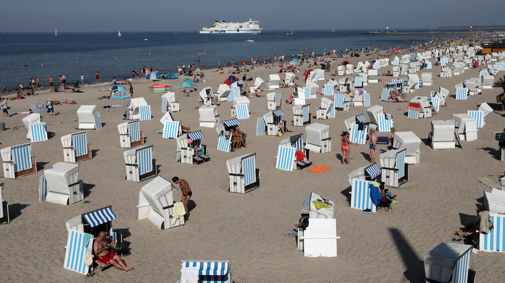 15.09.2020, Mecklenburg-Vorpommern, Warnemünde: Badegäste genießen das ungewöhnlich warme Spätsommerwetter am Ostseestrand, im Hintergrund läuft die Schweden-Fähre "Nils Holgersson" von TT-Line ein. Die September-Hitze sorgt in Deutschland für Temper