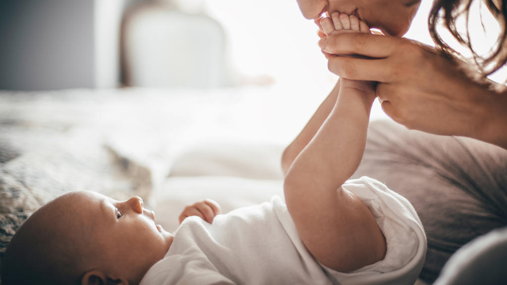 A happy mother kisses her baby's feet while she is lying down. The baby is looking up at her wearing a white baby grow.