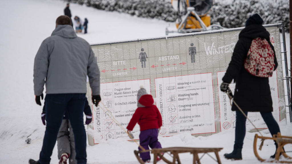 01.01.2021, Nordrhein-Westfalen, Winterberg: Ausflüger mit Schlitten gehen an einer geschlossenen Skipiste an einem Banner mit Corona-Verhaltensregeln vorbei. Die Stadt Winterberg bittet wegen der Corona-Pandemie auf Besuche in dem Wintersportort und