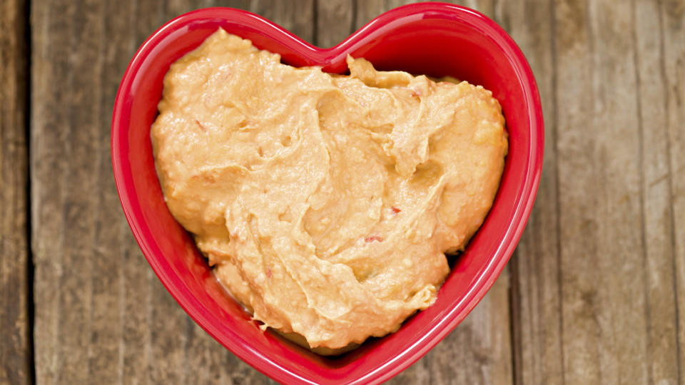 An overhead shot of a red heart shaped bowl with heart healthy rosasted red pepper humus. Shot on a grungy old wooden table. Humus is a Mediterranean spread made out of chickpeas or garbanzo beans.