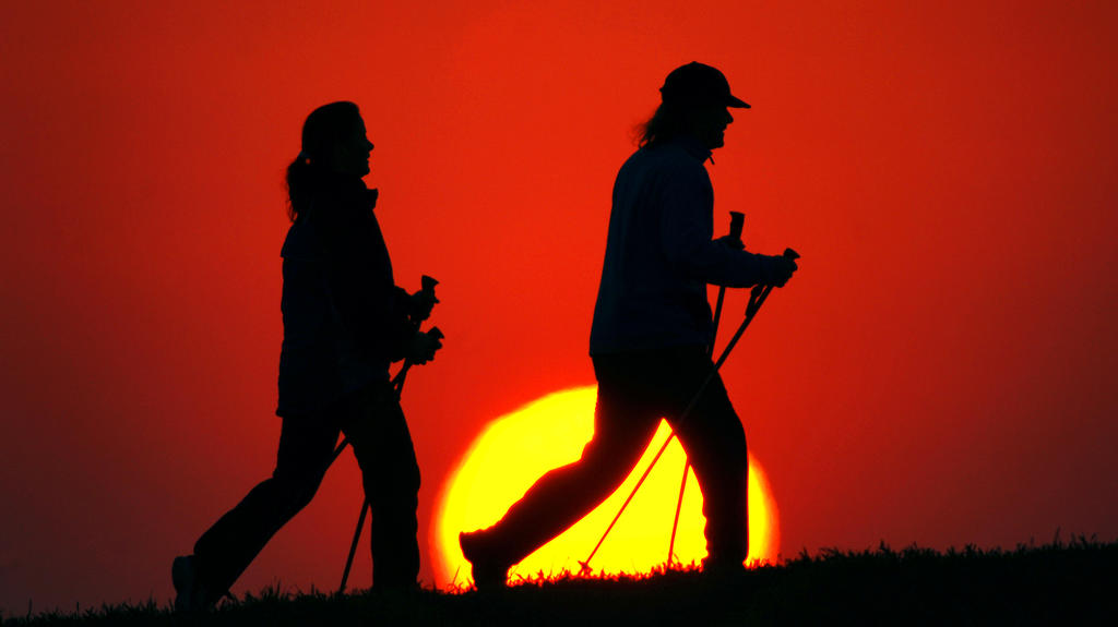 Bei Sonnenuntergang auf einem Feld sieht man zwei Personen die Sportart Nordic Walking, erkennbar an den Gehstöcken, ausüben.