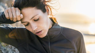 A photo of tired sporty woman looking down. Exhausted female athlete is wiping her forehead. She is wearing sportswear at beach.