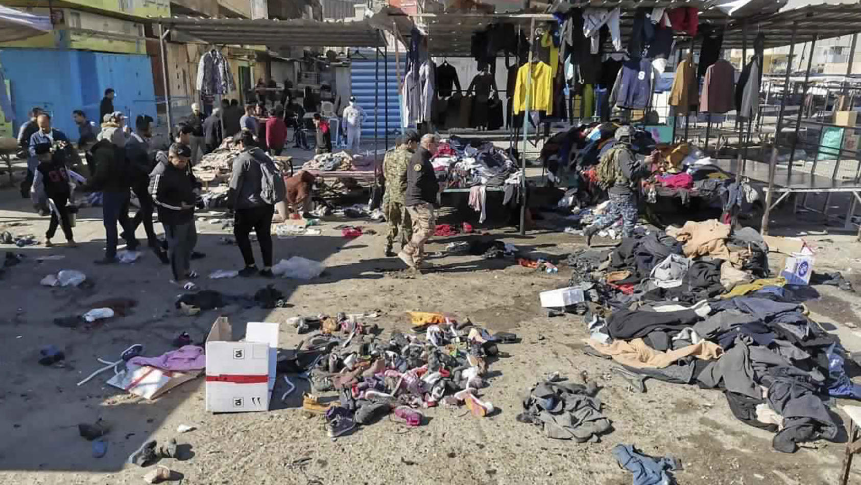 21.01.2021, Irak, Bagdad: Menschen und Sicherheitskräfte gehen in der irakischen Hauptstadt am Ort eines tödlichen Anschlags, einem Markt, auf dem gebrauchte Kleidung verkauft wurde, an auf dem Boden verstreuten Kleidung und Schuhen vorbei. Bei zwei 
