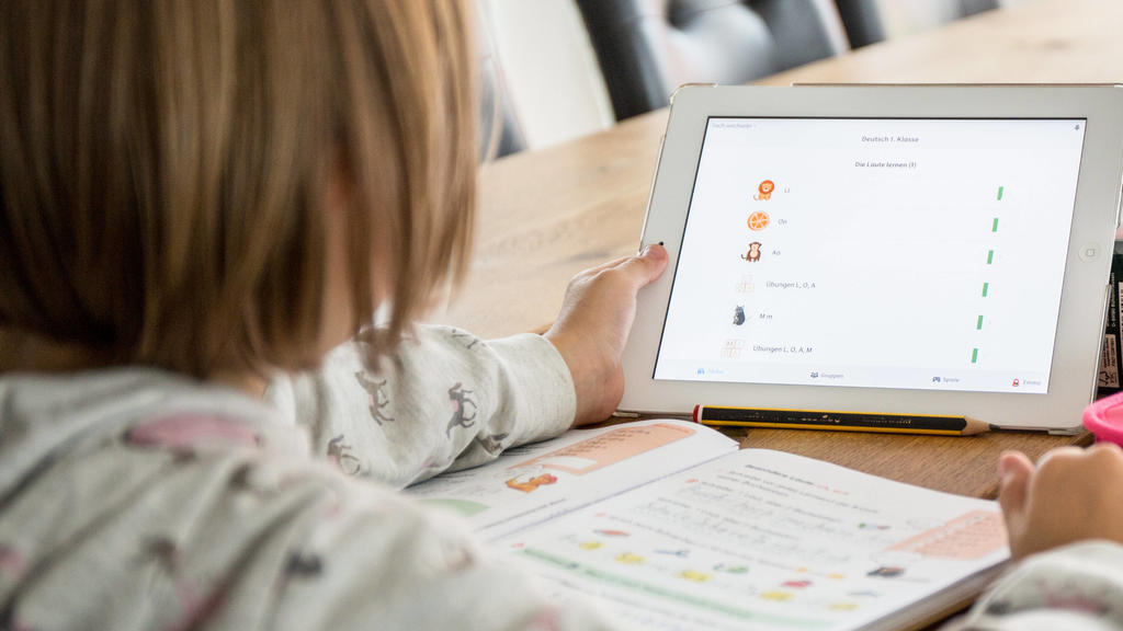 Ein Schulkind macht zu Hause am Tablet mit einem Online-Lernprogramm Homeschooling. Vor dem Kind liegt ein aufgeschlagenes Schulbuch und ein Bleistif