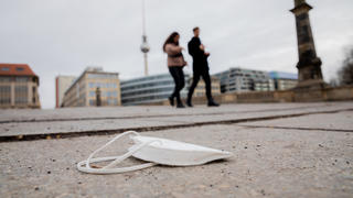 20.01.2021, Berlin: Eine FFP2-Maske liegt auf der Friedrichsbrücke, während im Hintergrund Passanten vorbeigehen. Foto: Christoph Soeder/dpa +++ dpa-Bildfunk +++