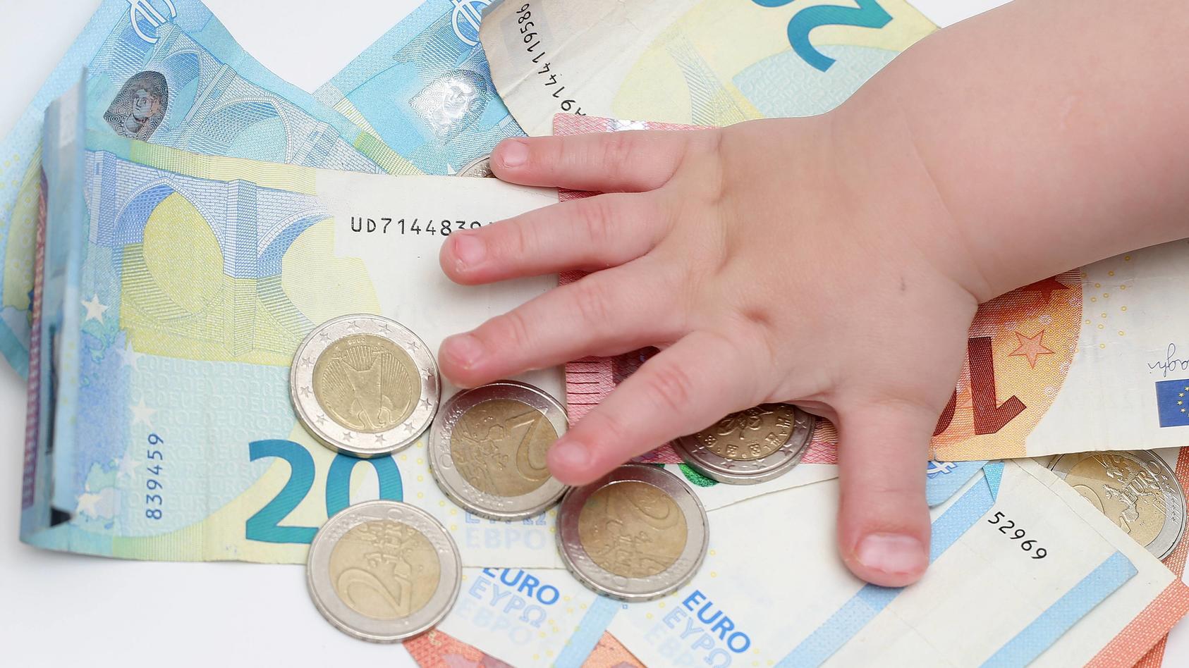Kinderhand greift nach Stapel von 2 Euro Muenzen auf Geldscheinen