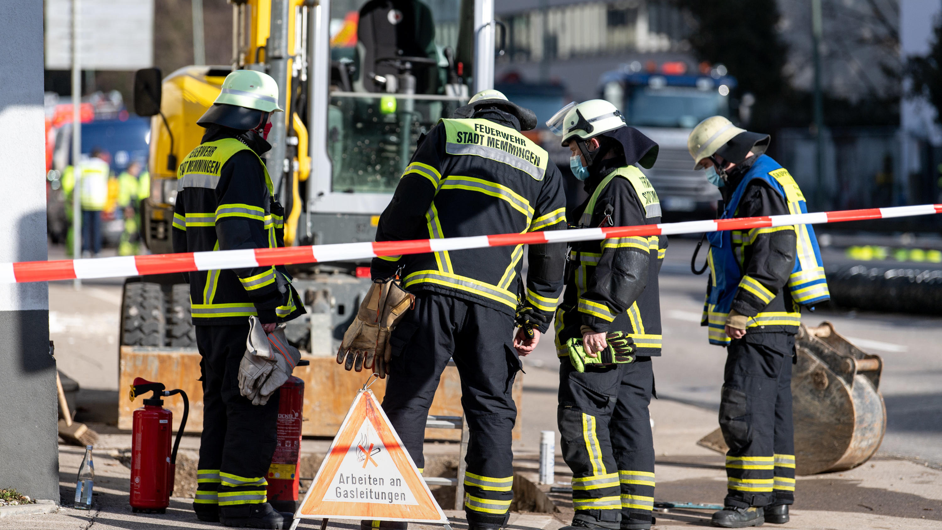 05.02.2021, Bayern, Memmingen: Einsatzkräfte der Feuerwehr stehen hinter einem Schild mit der Aufschrift «Arbeiten an Gasanlagen» an einer geöffneten Gehwegstelle vor einer Rettungswache vom Bayerischen Roten Kreuz, in der es zu einer Explosion gekom