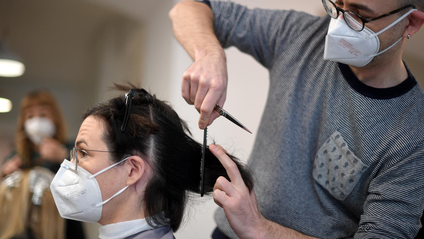 Ein Friseur schneidet einer Kundin in seinem Salon die Haare; beide tragen FFP2-Masken.
