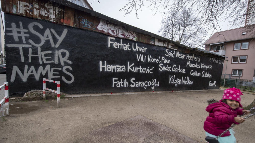 ARCHIV - 02.03.2020, Hessen, Hanau: Ein kleines Mädchen schaukelt vor der Wand eines Jugendzentrums, auf dem die Namen der Opfer des Anschlags vom 19. Februar geschrieben stehen. Links steht der Hashtag #Saytheirnames ("Nennt ihre Namen"), der Name d