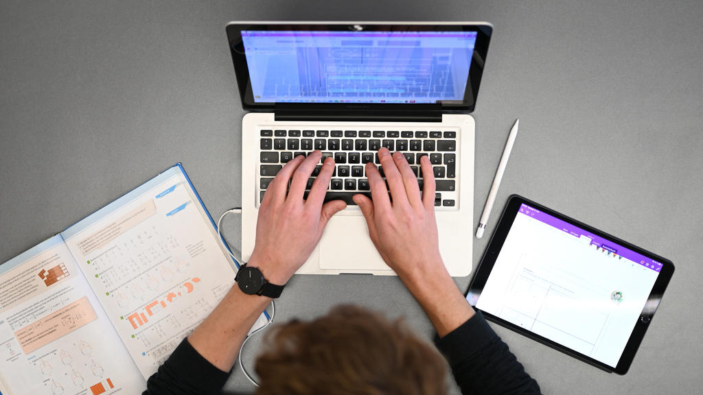 Ein Schüler sitzt vor seinem Laptop, neben ihm liebt ein Schulbuch und ein Tablet. Er befindet sich offenbar im Fernunterricht.