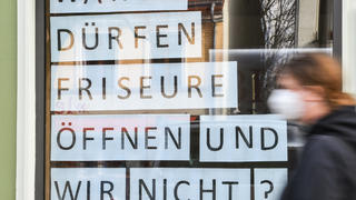 19.02.2021, Berlin: An dem Schaufenster eines Bekleidungsgeschäfts in Berlin Friedrichshagen steht die Frage ·Warum dürfen Friseure öffnen und wir nicht?·. Davor läuft eine Person mit Mund-Nasen-Bedeckung. Foto: Kira Hofmann/dpa-Zentralbild/dpa +++ dpa-Bildfunk +++