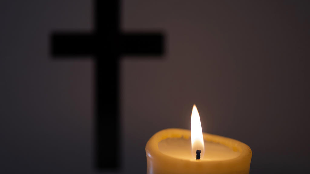 Im Vordergrund des Bildes brennt eine Kerze, im Hintergrund sieht man ein Kreuz.