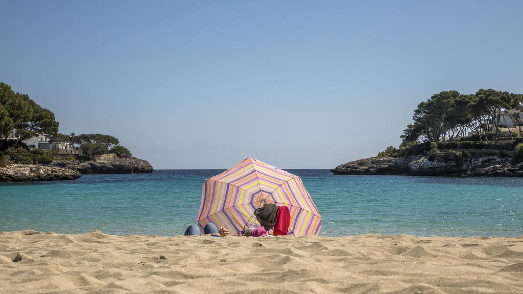 dpatopbilder - 25.03.2021, Spanien, Cala D'or: Zwei Personen liegen unter einem Sonnenschirm an einem Strand auf Mallorca. Angesichts der Corona-Pandemie sollen sich Flugpassagiere vor dem Start nach Deutschland künftig generell testen lassen müssen