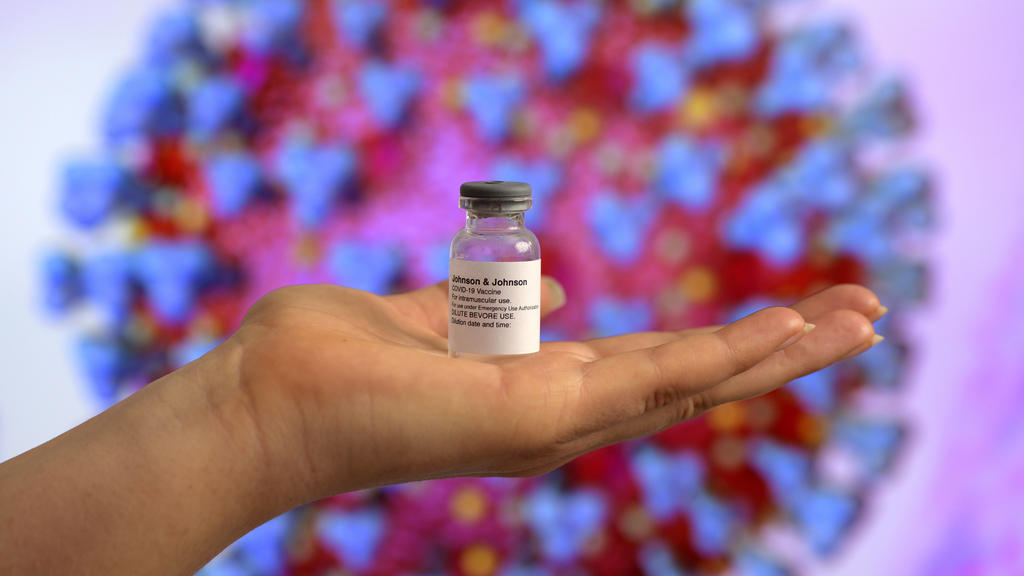 Symbolbild: Zugelassene Impfstoffe von Johnson & Johnson wird auf einer ausgestreckten Hand präsentiert, im Hintergrund sieht man ein Symbolfoto des Coronavirus.