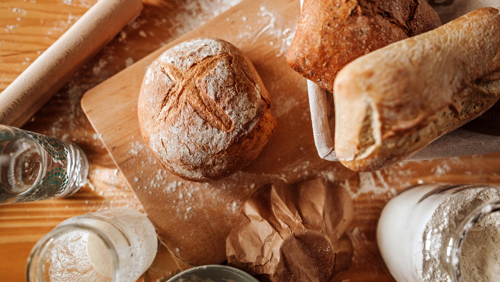 Auf einem Brettchen auf einem Esstisch liegen verschiedene Utensilien, unter anderem ein frisch gebackenes Sauerteigbrot.