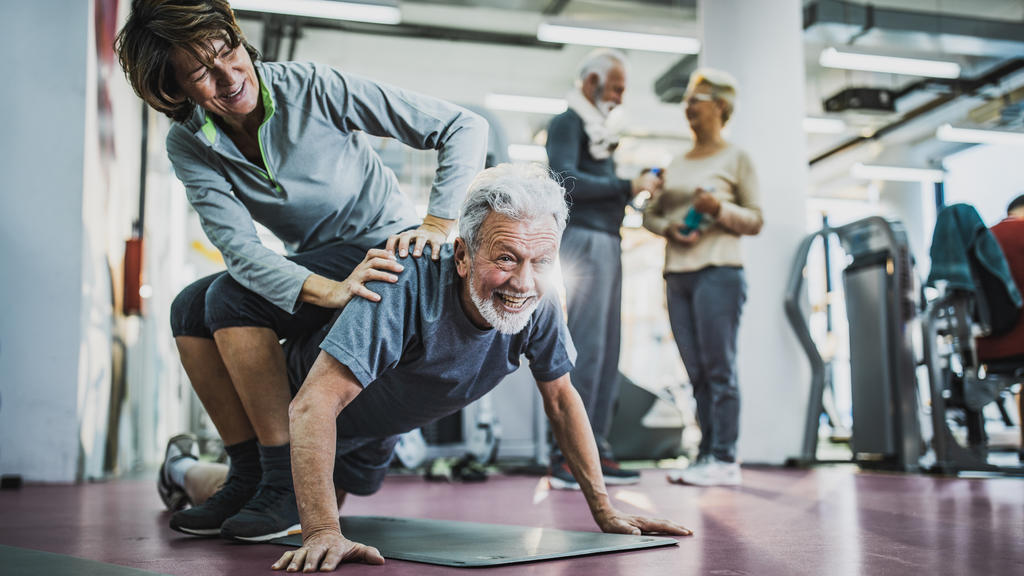 Ein glücklich wirkendes Paar hat Spaß beim Workout im Fitnessstudio, während der Mann eine Liegestütze durchführt.