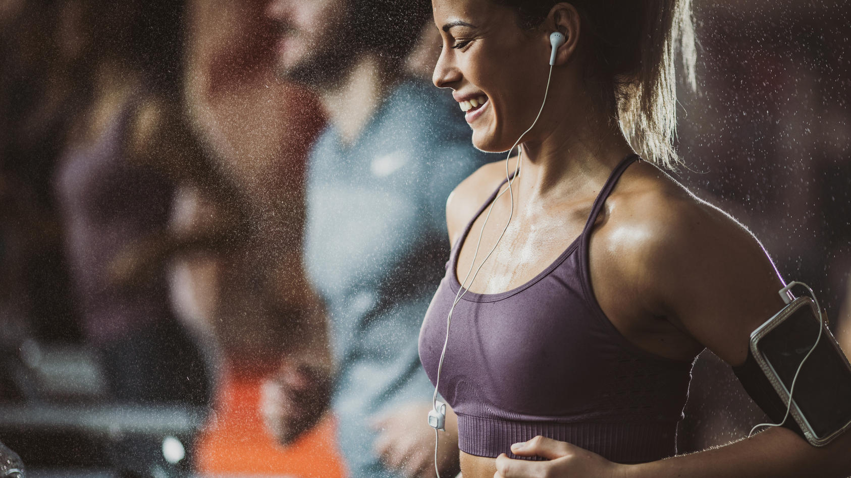 Eine glücklich aussehende Frau trainiert auf dem Laufband im Fitnessstudio. Über Kopfhörer hört sie Musik auf ihrem Handy.