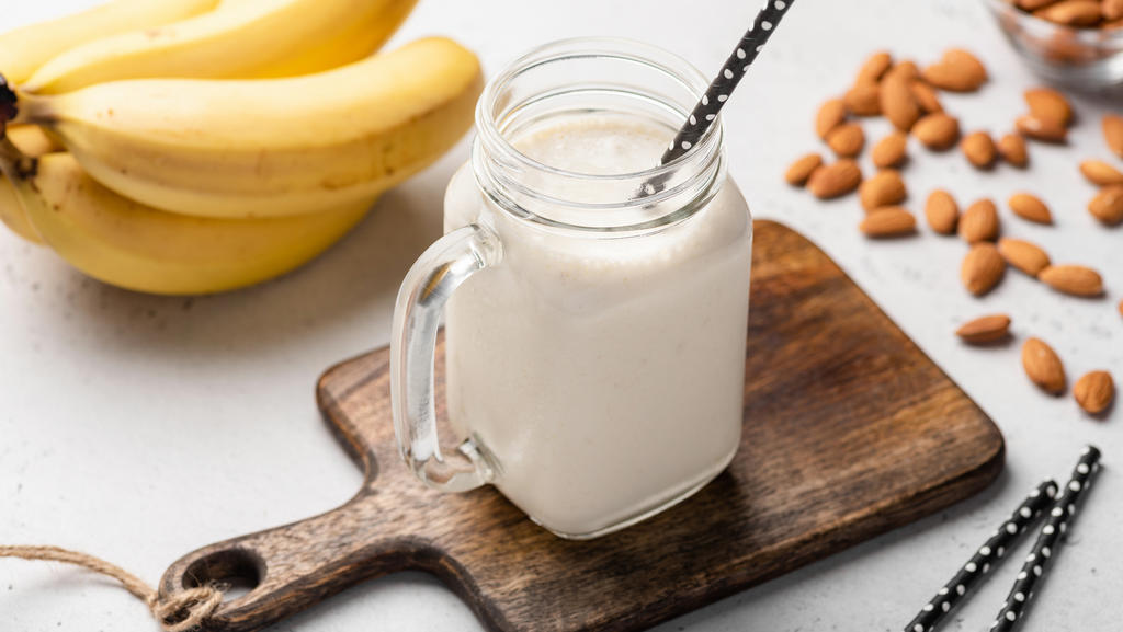 Ein leckerer Bananen-Protein-Smoothie ist hübsch angerichtet auf einem kleinen Holzbrettchen. Daneben liegen Strohalme, Bananen und Mandeln.