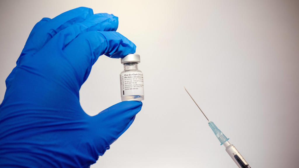 Eine Ampulle mit dem Impfstoff gegen Covid-19 von Pfizer-BioNTech wird in der linken Hand gehalten und die Spritze mit dem aufgezogenen Impfstoff sieht man daneben.