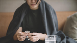 Frau mit Decke über den Schultern sitzt vor Glas und Schmerztablette