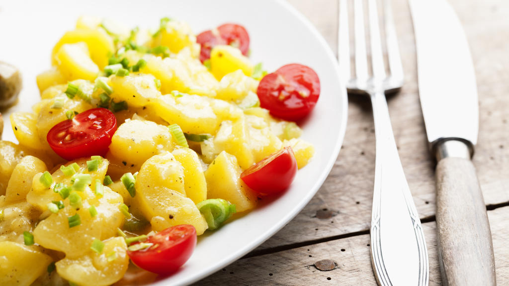 Eine Portion Kartoffelsalat liegt auf einem Teller, daneben sieht man Besteck liegen.