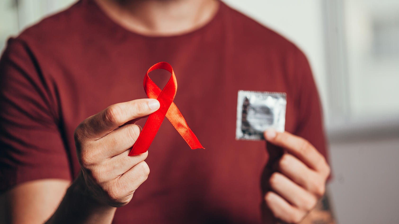 Ein Mann hält in der einen Hand ein Kondom, in der anderen eine rote Schleife, die für die Aufklärung der Krankheit HIV steht.