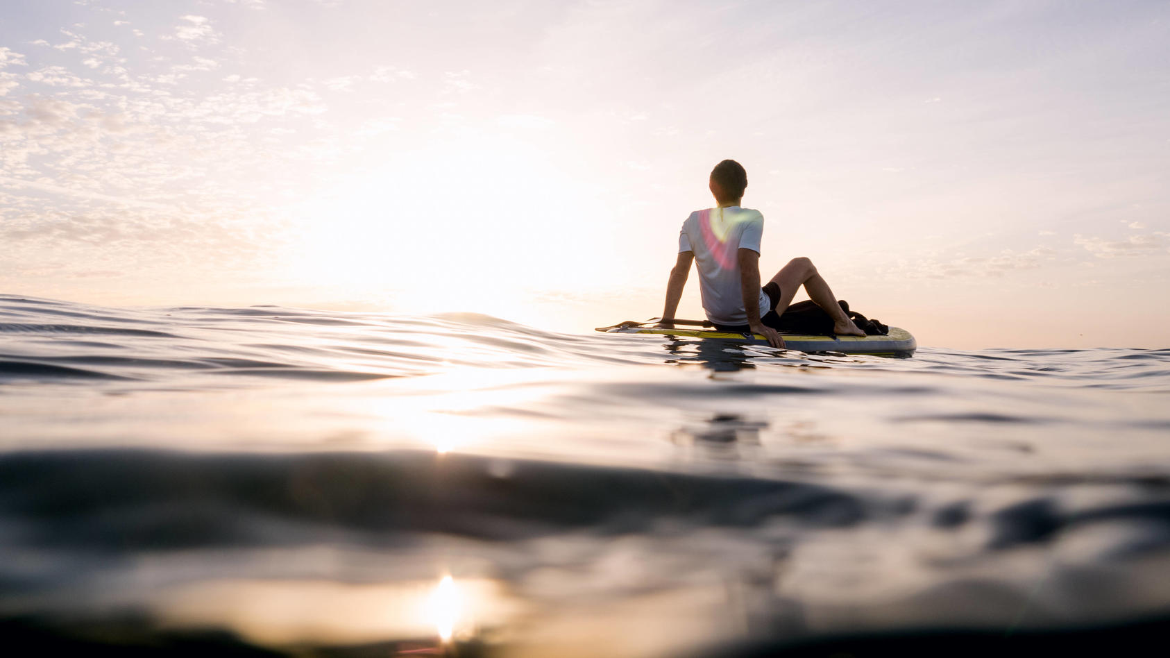 Auf dem offenen Meer entspannt ein Mann auf einem Surfboard, man sieht im Hintergrund wie die Sonne untergeht.