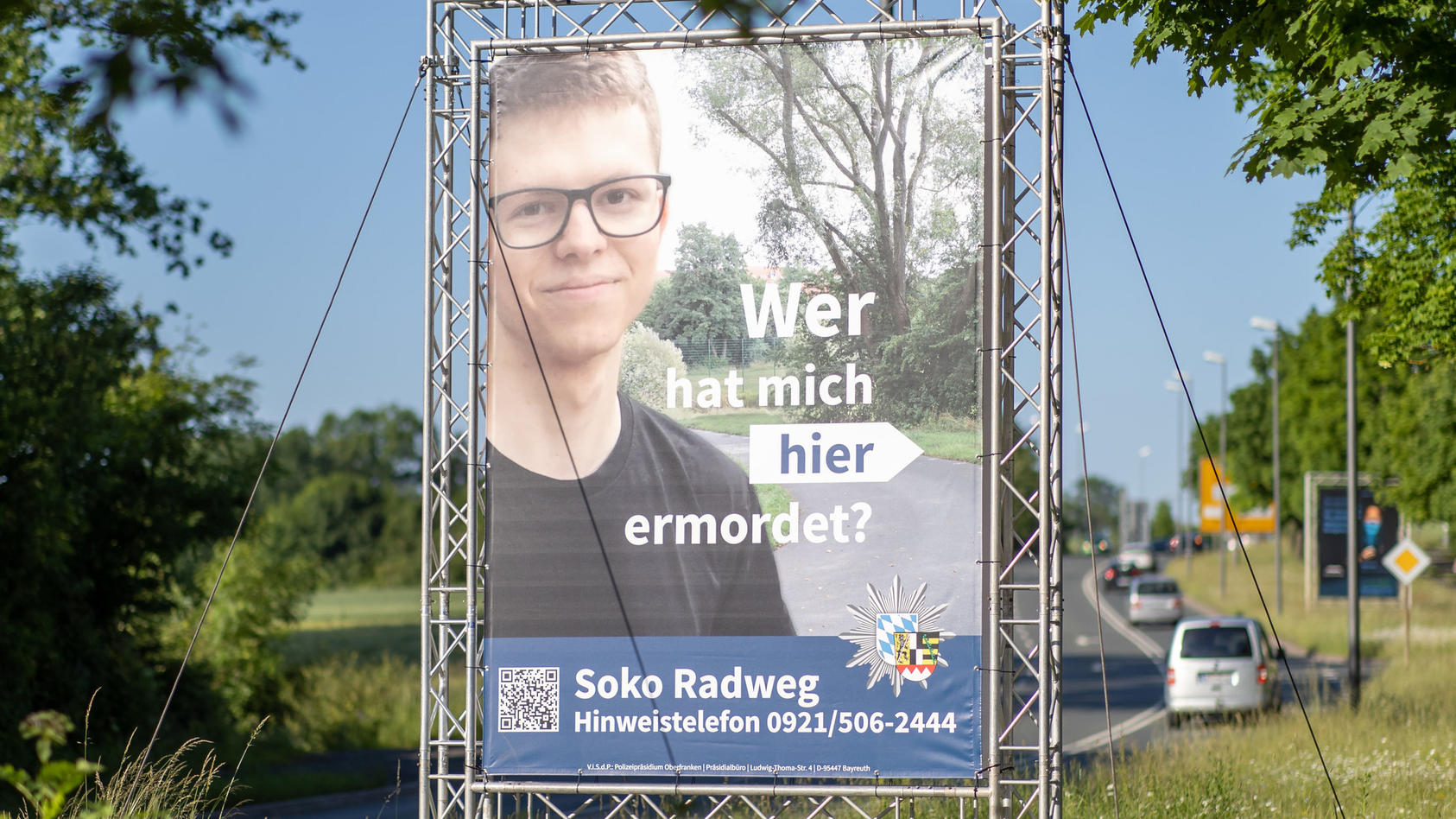 14.06.2021, Bayern, Bayreuth: "Wer hat mich hier ermordet?" steht auf einem großen Plakat mit Foto des Opfers neben dem Tatort. Mithilfe des Plakats sucht die Polizei nach einem Täter, der im Sommer 2020 einen 24-Jährigen in Bayreuth umgebracht haben