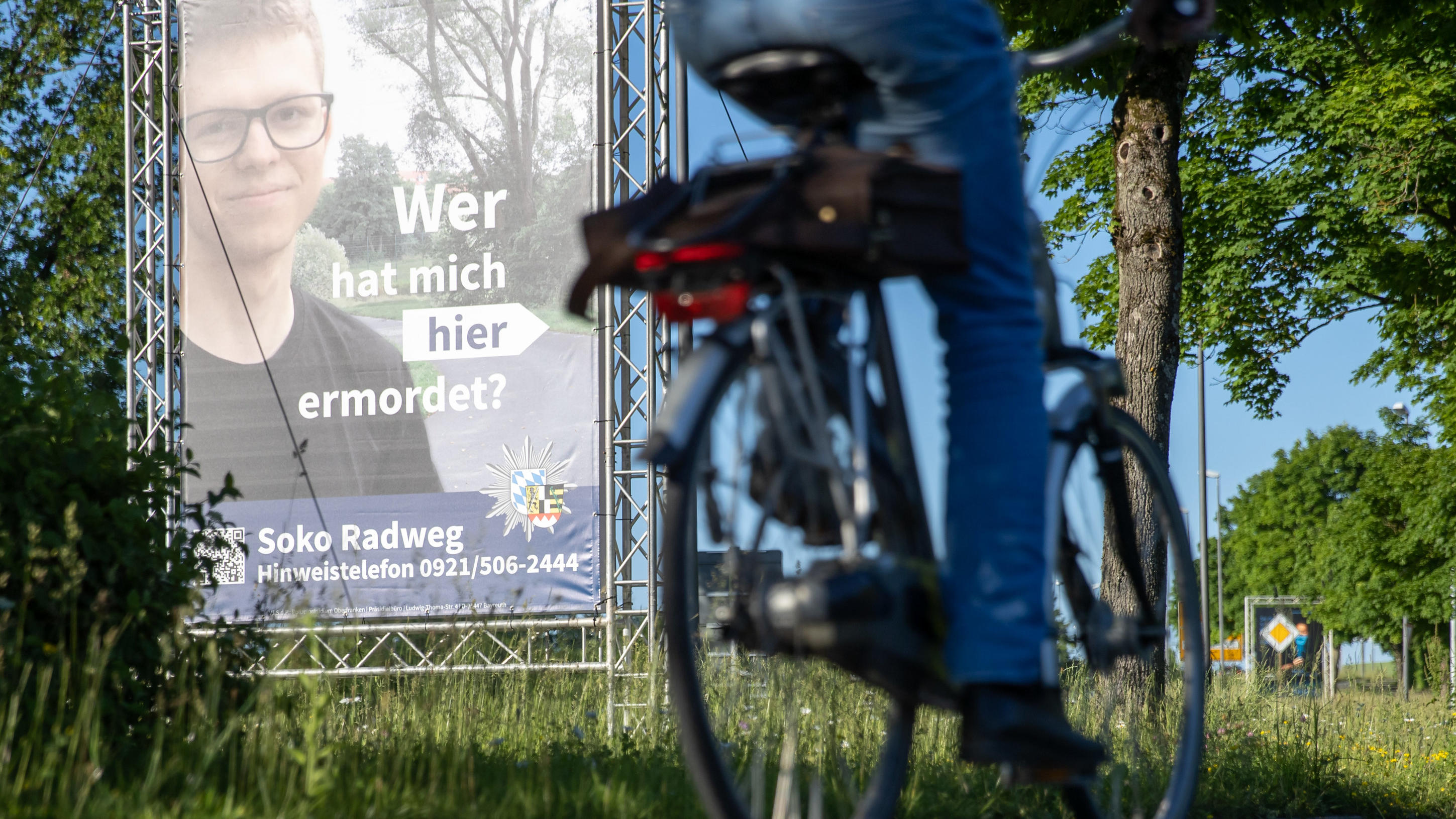 14.06.2021, Bayern, Bayreuth: "Wer hat mich hier ermordet?" steht auf einem großen Plakat mit Foto des Opfers neben dem Tatort. Mithilfe des Plakats sucht die Polizei nach einem Täter, der im Sommer 2020 einen 24-Jährigen in Bayreuth umgebracht haben